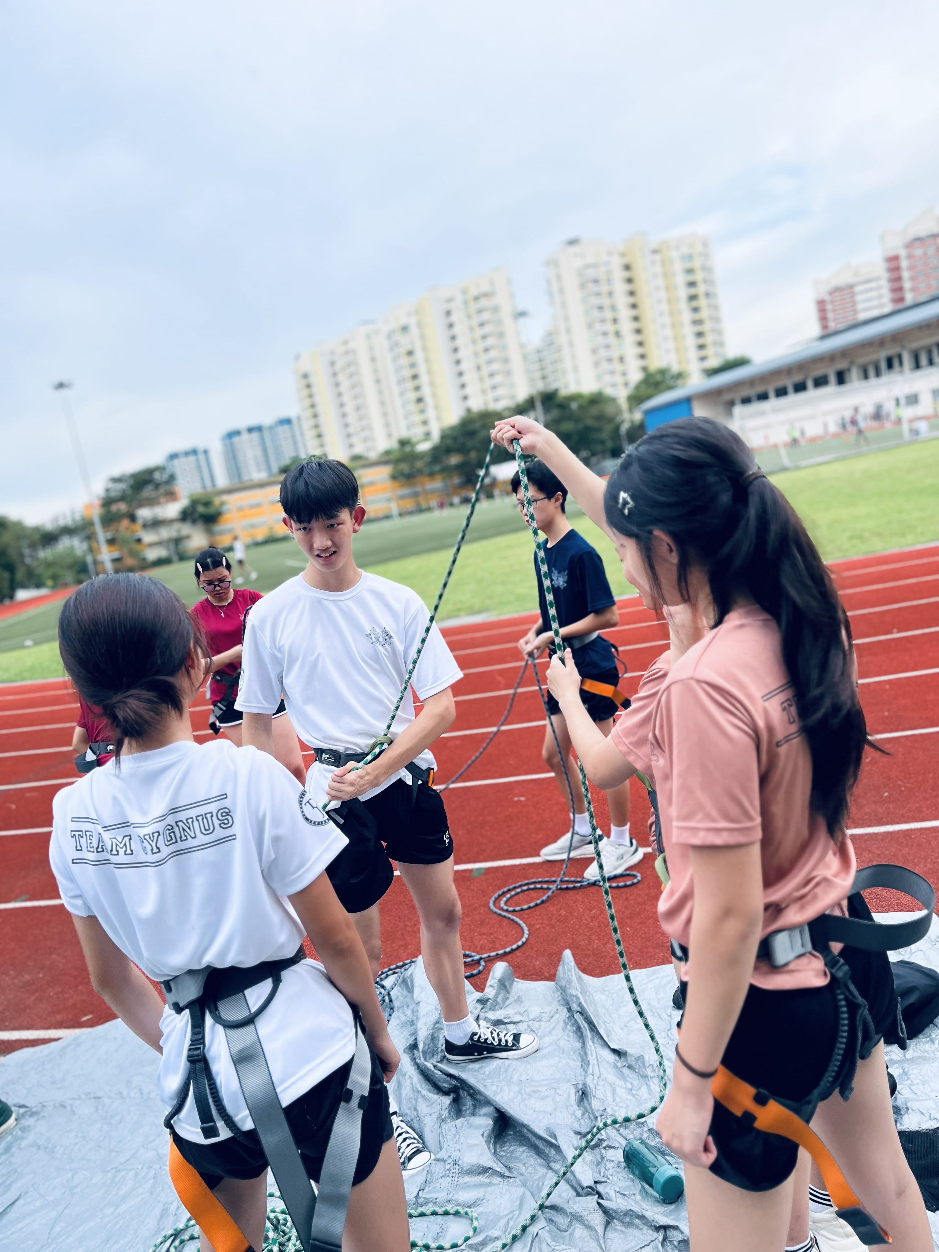 Students in climbing harnesses practise rope handling on a running track, with HDB flats visible in the background.
